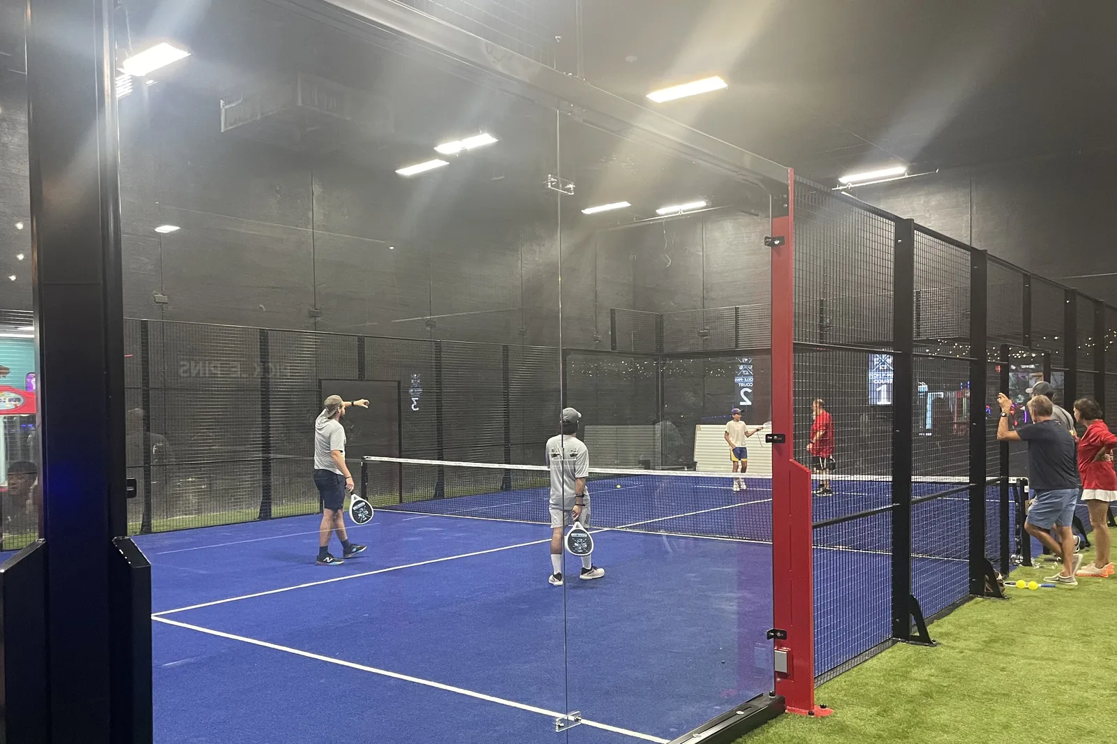 Doubles play on an indoor panoramic padel court with glass walls and a red corner post, inside a converted building in New Orleans, Louisiana